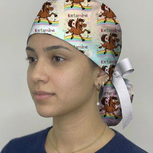 May include: A scrub cap featuring a repeating cartoon horse pattern on a light background with rainbows, flowers, and the word ketamine printed in colorful letters. The cap includes a white ribbon tie at the back to secure the fit.