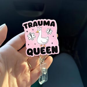 May include: A pink badge reel with the words "TRAUMA QUEEN" in black, featuring a cartoon goose wearing a crown and medical symbols. The reel has a clear plastic clip and a metal retractable cord.