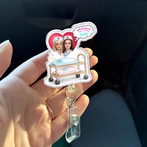 May include: A glittery acrylic badge reel featuring two cartoon nurses in a heart-shaped frame above a medical cart with two IV bags. A speech bubble above them contains the text: HOP IN BESTIE we're starting GRRT. The design has a silver glitter border.