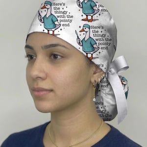 Pu&ograve; includere: Una cuffia chirurgica bianca con un motivo ripetuto di un'anatra dei cartoni animati che indossa una casacca chirurgica color ottanio e tiene in mano un bisturi. Il testo "Here's the thingy with the pointy end" &egrave; stampato accanto a ogni anatra.