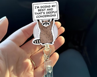 May include: A retractable badge reel featuring a cartoon raccoon holding a sign. The sign reads "I'M DOING MY BEST AND THAT'S DEEPLY CONCERNING". The reel has a clear plastic clip and a silver-colored metal clasp.