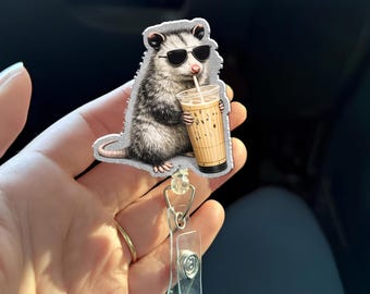 May include: A badge reel featuring a cartoon opossum wearing sunglasses and drinking iced coffee. The opossum is gray and white, and the coffee cup is tan with a clear lid. The reel has a clear plastic clip and a retractable cord.
