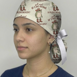 May include: A cream-colored scrub cap featuring a repeating pattern of green frogs wearing brown cowboy hats and boots. The text "ya'll mind IF I DO SOME LOLLYGAGGIN?" is printed in brown lettering across the fabric. A white ribbon is tied at the back.