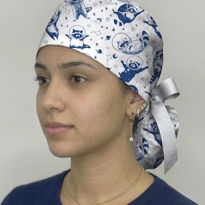 May include: A white scrub cap featuring a repeating blue pattern of raccoons wearing space helmets, surrounded by stars and planets. The cap has a gathered back with a light gray ribbon tie closure, providing a secure and adjustable fit.