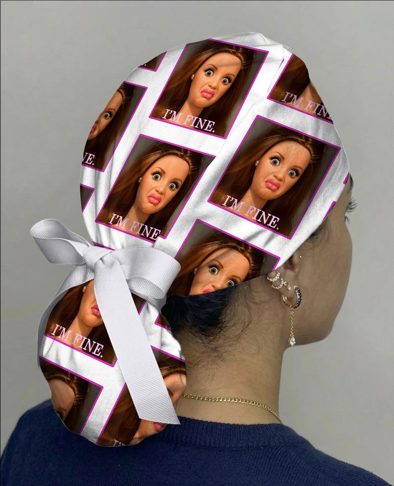 Peut inclure: Une charlotte de bloc op&eacute;ratoire blanche avec un motif r&eacute;p&eacute;titif repr&eacute;sentant un visage de poup&eacute;e &agrave; l'expression contrari&eacute;e et le texte I'M FINE. imprim&eacute; en blanc. Le bonnet comporte un ruban blanc &agrave; l'arri&egrave;re pour ajuster la taille.