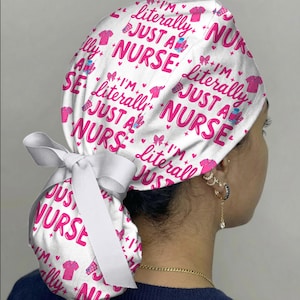 May include: White scrub cap with a pink and white pattern. The cap features the text "I'm Literally Just a Nurse" in pink, along with pink hearts, bows, and medical-themed graphics. A white ribbon is tied at the back.