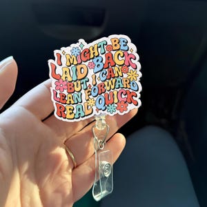 Puede incluir: Un portatarjetas de colores con el texto "I MIGHT BE LAID BACK BUT I CAN LEAN FORWARD REAL QUICK" en una fuente retro. El portatarjetas tiene un soporte de plástico transparente y un cordón retráctil.