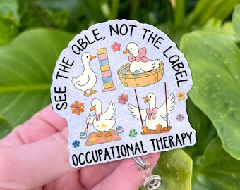 Könnte beinhalten: Ein Abzeichen mit dem Text "SEE THE ABLE, NOT THE LABEL" und "OCCUPATIONAL THERAPY". Das Abzeichen zeigt Cartoon-Enten, die sich mit verschiedenen Aktivitäten beschäftigen, darunter eine Schaukel, Malerei und ein Korb. Das Abzeichen wird vor einem Hintergrund aus grünen Blättern gehalten.