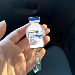 May include: A badge reel with a white and blue DIPRIVAN propofol vial design. The vial has a blue cap and text including "10mg/mL propofol" and "FOR I.V. ADMINISTRATION". The reel has a clear plastic clip and a retractable cord.
