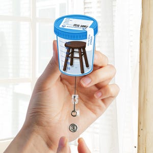 May include: A novelty badge reel featuring a cartoon image of a small wooden stool inside a blue-lidded specimen container. The container has the words "STOOL SAMPLE" and "BIOHAZARDOUS" printed on it. The reel has a clear plastic strap and a metal snap.