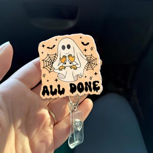 Puede incluir: Un portatarjetas con temática de Halloween con un fantasma de dibujos animados, manos naranjas y una sábana blanca. El portatarjetas tiene las palabras "ALL DONE" en letras negras, con telarañas y murciélagos. El portatarjetas está sujeto a un clip de plástico transparente.