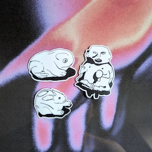 May include: Three black and white line drawings of a frog, a rabbit, and a person. The frog is sitting on its hind legs with its front legs outstretched. The rabbit is sitting on its hind legs with its front legs tucked under its chin. The person is standing with their arms crossed and has a large head with a simple face.