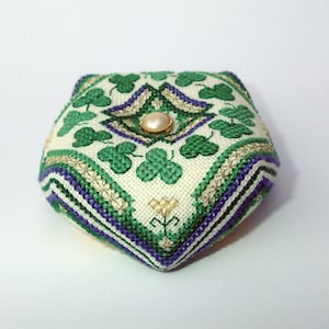 Könnte beinhalten: Ein weißer Stoff-Nadelkissen mit einem grünen Kleeblatt-Muster und einem goldenen und violetten Besatz. Ein Perlknopf befindet sich in der Mitte des Nadelkissens.