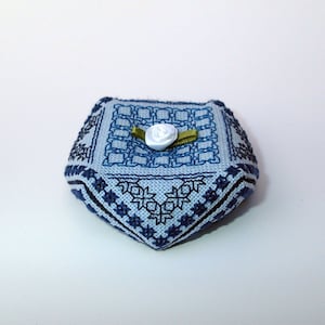 May include: A small, square, blue and white needlepoint cushion with a white satin bow on top. The cushion has a geometric pattern and a black border.
