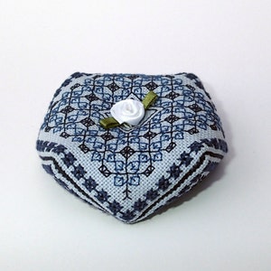May include: A small, heart-shaped pincushion with a blue and white geometric pattern. A white satin rose with a green ribbon is centered on the pincushion.