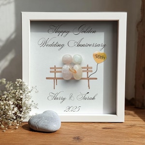 Könnte beinhalten: Ein gerahmtes Kunstwerk zur Feier einer goldenen Hochzeit. Das Kunstwerk zeigt eine Bank mit Kieselsteinfiguren, einen 50. Jahrestag-Ballon und die Namen "Harry & Sarah" mit dem Jahr 2025. Ein herzförmiger Stein liegt davor.