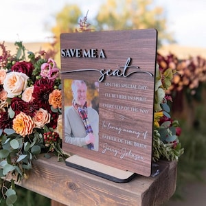 Op de afbeelding: Een houten herdenkingsbord met de woorden "SAVE ME A seat" in zilveren letters. Het bord bevat een foto van een man en de tekst "In loving memory of FATHER OF THE BRIDE Jerry Johnson."