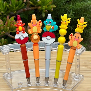 Peut inclure: Cinq stylos colorés avec différents personnages de Pokemon. Les stylos sont présentés dans un porte-stylos en plastique transparent sur une surface en bois.