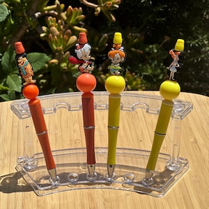 Puede incluir: Cuatro bolígrafos con figuras de personajes de dibujos animados, expuestos en un soporte de plástico transparente. Los bolígrafos tienen barriles naranjas y amarillos, con detalles plateados. Las figuras representan personajes de una serie animada clásica.