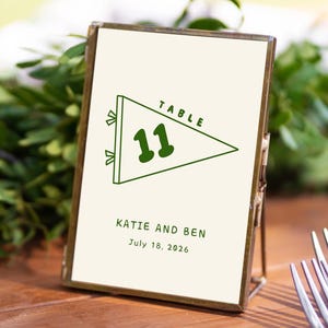 May include: A table number card with a green pennant design and the number 11. The card also includes the names "Katie and Ben" and the date "July 18, 2026". The card is in a gold frame.