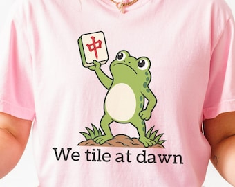 May include: A light pink t-shirt featuring a green frog standing on a small mound of dirt, holding a white mahjong tile with a red character. Below the frog, the text reads "We tile at dawn" in a dark, simple, sans-serif font.
