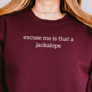 May include: A maroon long-sleeve crewneck sweatshirt featuring the white text "excuse me is that a jackalope" printed on the center chest. The garment has a ribbed neckline, cuffs, and hem, and is worn with blue denim jeans.