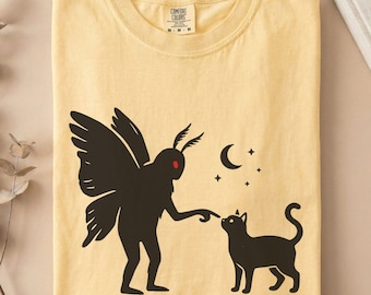 May include: A butter yellow t-shirt featuring a black graphic of a moth-like creature with red eyes pointing at a black cat. Above the cat are a crescent moon and small stars. The shirt has a Comfort Colors brand label at the inner neckline.