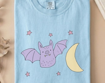 May include: A light blue t-shirt featuring a graphic of a purple bat with small fangs, a yellow crescent moon, and several small pink stars. The shirt has a crew neckline and a Comfort Colors brand label on the inside collar with the text Chambray.