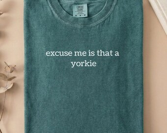 May include: A blue spruce coloured short-sleeve t-shirt with a crew neckline. The centre of the shirt features the white text, excuse me is that a yorkie. The garment includes a Comfort Colors brand label on the inside of the collar.