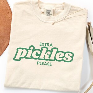 May include: Ivory-colored t-shirt with the text "EXTRA pickles PLEASE" in green. The shirt has a crew neck and a small tag on the upper back. A gold necklace and part of a brown hat are visible.