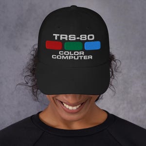 Op de afbeelding: Een zwarte baseballpet met een wit geborduurd logo met de tekst "TRS-80 COLOR COMPUTER". Het logo bevat ook drie gekleurde vierkanten: rood, groen en blauw.