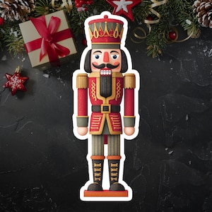 Puede incluir: Imagen estilo pegatina de un soldado cascanueces tradicional. El cascanueces viste un uniforme rojo con detalles dorados, barba negra y corona. El fondo incluye adornos navideños, una caja de regalo y ramas de pino.