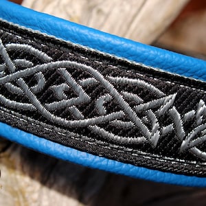 May include: A close-up of a blue leather dog collar with a black and silver Celtic knot design. The collar is made of high-quality leather and features a durable, stylish design.