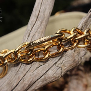 Puede incluir: Un collar de perro de cadena dorada con una tela estampada en negro y dorado. La cadena tiene un cierre con las palabras "Made in Germany" grabadas.