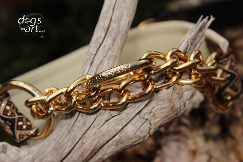 Puede incluir: Un collar de perro de cadena dorada con una tela estampada en negro y dorado. La cadena tiene un cierre con las palabras "Made in Germany" grabadas.