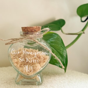 Pode incluir: Um frasco de vidro em forma de coração com tampa de cortiça cheio de areia. O frasco está amarrado com um barbante e tem o texto "Our Honeymoon St. Kitts 2025" escrito nele.