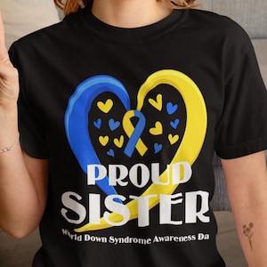 May include: Black t-shirt with a yellow and blue heart design and the text "Proud Sister" and "World Down Syndrome Awareness Day".