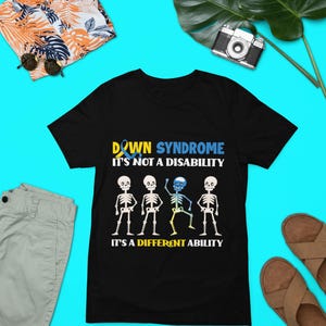 May include: Black t-shirt featuring a graphic of four skeletons, one with a blue and yellow ribbon. The text reads "Down Syndrome It's not a disability It's a different ability".