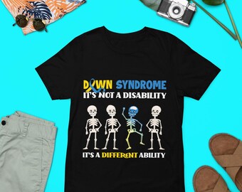Può includere: Maglietta nera con una grafica di quattro scheletri, uno con un nastro blu e giallo. Il testo recita "Sindrome di Down Non è una disabilità È una capacità diversa".