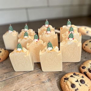 May include: A collection of rectangular, light beige soap bars with a marbled texture. Each bar is topped with a small, decorative figurine of a gnome wearing a green, white-spotted hat shaped like a Christmas tree. Several chocolate chip cookies.
