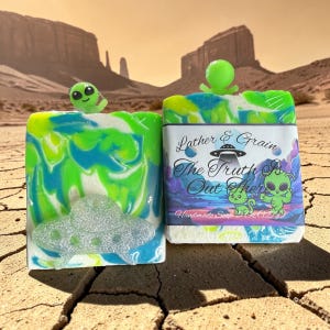 May include: Two handmade soaps with an alien theme. One soap has a green and blue swirl design with a glittery UFO. The other soap has a label that reads "Lather & Grain The Truth Is Out There". Each soap has a small alien figure on top.