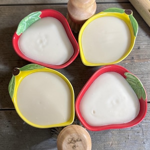 May include: Four fruit-shaped soap dishes, two red and two yellow, each filled with white soap. Two wooden handled scrub brushes are also in the image. The text "Lather & Grain" is on the top of the brushes.