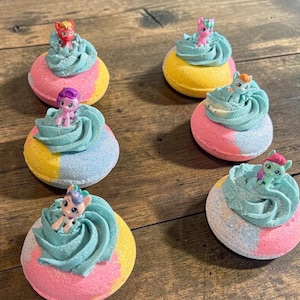 May include: Six colorful bath bombs with a cupcake design. Each bath bomb is split into pink, yellow, and blue sections, topped with a swirl of blue frosting and a small toy pony. The bath bombs are arranged on a wooden surface.