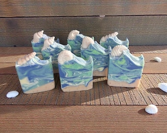 May include: Artisan soaps with a beach theme. Each bar features a wave design in shades of blue and green, topped with a seashell. The soaps have a light tan base, resembling sand. Several seashells are scattered around the soaps on a wooden surface.