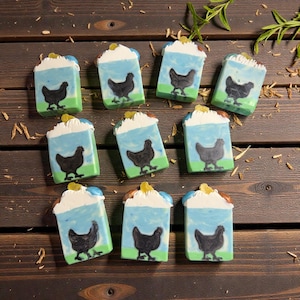 May include: A collection of handmade soaps with a whimsical design. Each rectangular bar features a black chicken silhouette against a blue sky background, with a green base and a white, textured top. The soaps are arranged on a dark wooden surface.