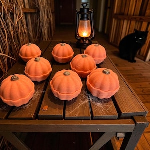 May include: A collection of eight orange pumpkin-shaped items with textured surfaces, arranged on a dark brown wooden table. Each pumpkin has a small, dark brown stem. A lit lantern and a black cat are in the background.