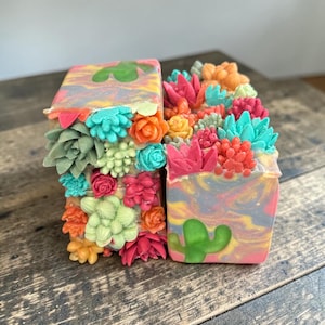 May include: Two rectangular bars of soap featuring a marbled base of pink, yellow, and blue swirls. The tops and sides are decorated with three-dimensional, colorful succulent and cactus shapes in shades of green, orange, pink, and teal.