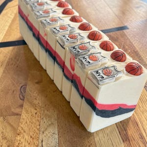 May include: A rectangular bar of handmade soap with a basketball theme. The soap has layers of white, red, and black. Each bar is topped with a small, orange basketball and a silver MVP emblem. The soap is displayed on a wooden surface.