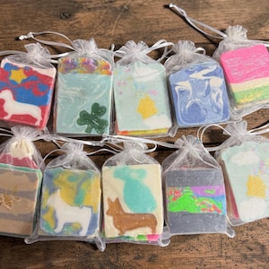 May include: A collection of ten decorative soaps, each individually packaged in a sheer organza bag. The soaps feature colorful designs, including a dachshund, shamrock, and various abstract patterns. The soaps are displayed on a wooden surface.