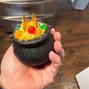 May include: A black cauldron filled with yellow material, gold coins, and colorful gems. The cauldron is held in a hand, and the background is a wooden surface and a stainless steel appliance.